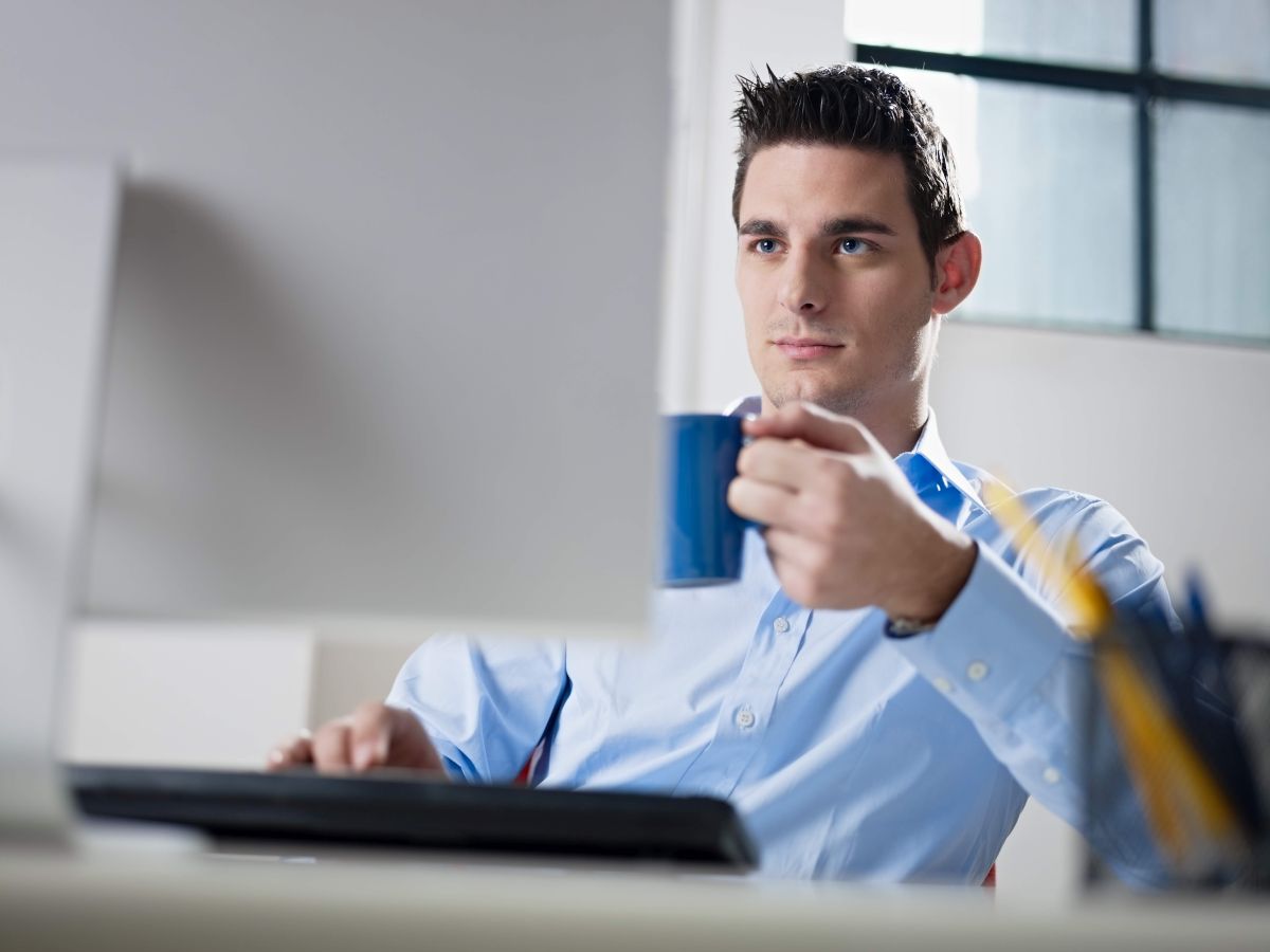 Opanowany i rozluźniony mężczyzna w niebieskiej koszuli patrzy na monitor z kubkiem w lewej ręce i prawą ręką na klawiaturze