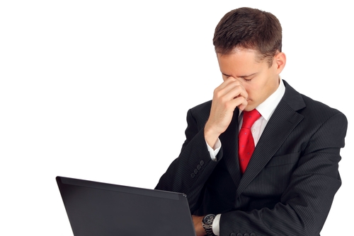 Mężczyzna w garniturze trzyma na kolanach laptop i chwyta się za nos ze spuszczoną głową
