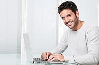 Szeroko uśmiechnięty brunet siedzi bokiem i pisze na laptopie