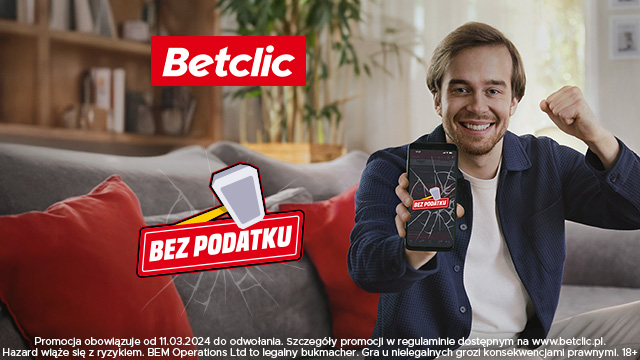Uradowany mężczyzna w garniturze i koszulce na kanapie na tle salonu z książkami pokazuje aplikację Betclic w telefonie i czerwony napis Betclic bez podatku