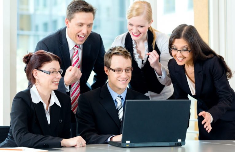 Seorang pria dalam setelan bisnis duduk di depan laptop dikelilingi oleh karyawan yang bahagia