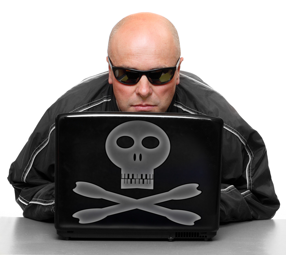 Łysy mężczyzna w ciemnych okularach siedzi za laptopem na którego obudowie jest trupia czaszka z piszczelami