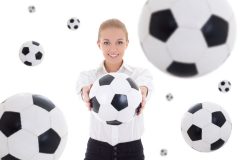 Uśmiechnięta kobieta w białej koszuli trzyma przed sobą piłkę nożną w otoczeniu wiszących piłek wokół