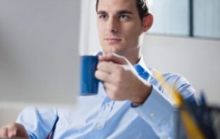 Opanowany i rozluźniony mężczyzna w niebieskiej koszuli patrzy na monitor z kubkiem w lewej ręce i prawą ręką na klawiaturze
