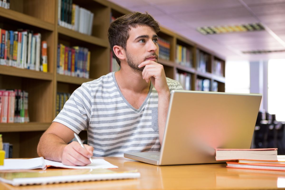 Mężczyzna w pasiastym podkoszulku siedzi przed laptopem i zastanawia się z dłonią przygotowaną do pisania w zeszycie na tle regału z książkami