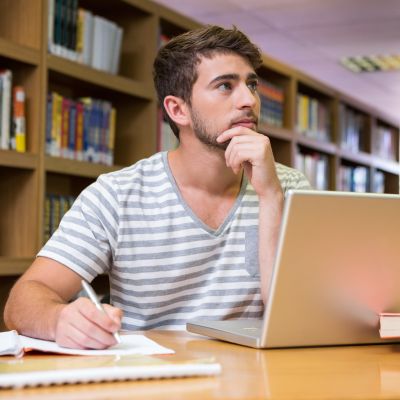 Mężczyzna w pasiastym podkoszulku siedzi przed laptopem i zastanawia się z dłonią przygotowaną do pisania w zeszycie na tle regału z książkami