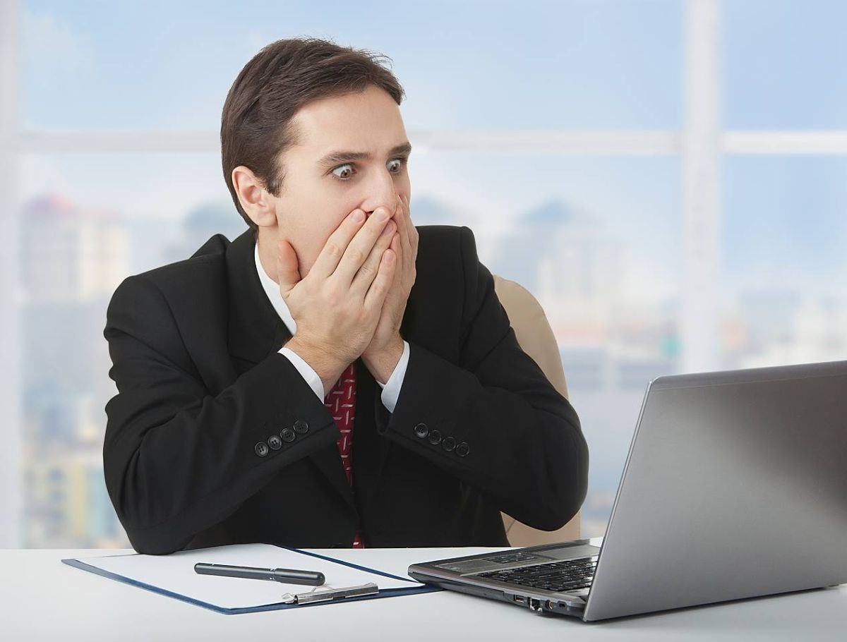 Przerażony mężczyzna w garniturze siedzi w biurze przy biurku przed laptopem i zakrywa sobie usta obiema rękami