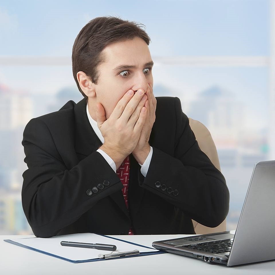 Przerażony mężczyzna w garniturze siedzi w biurze przy biurku przed laptopem i zakrywa sobie usta obiema rękami