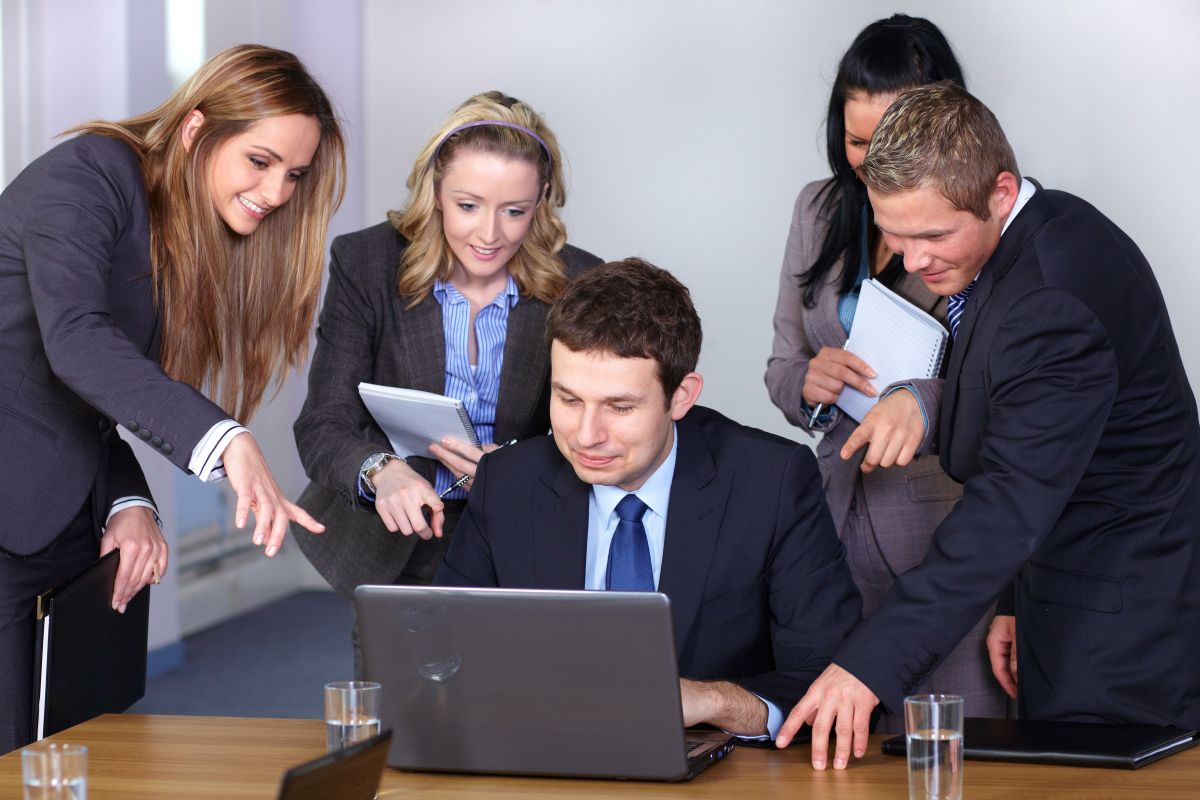Biznesman siedzący przed laptopem otoczony kolegami z biura