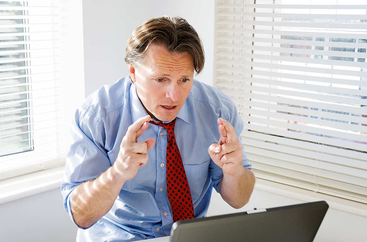 Stojący mężczyzna w niebieskiej koszuli i poluzowanym krawacie patrzy z niepokojem na laptopa ze splecionymi palcami obu dłoni