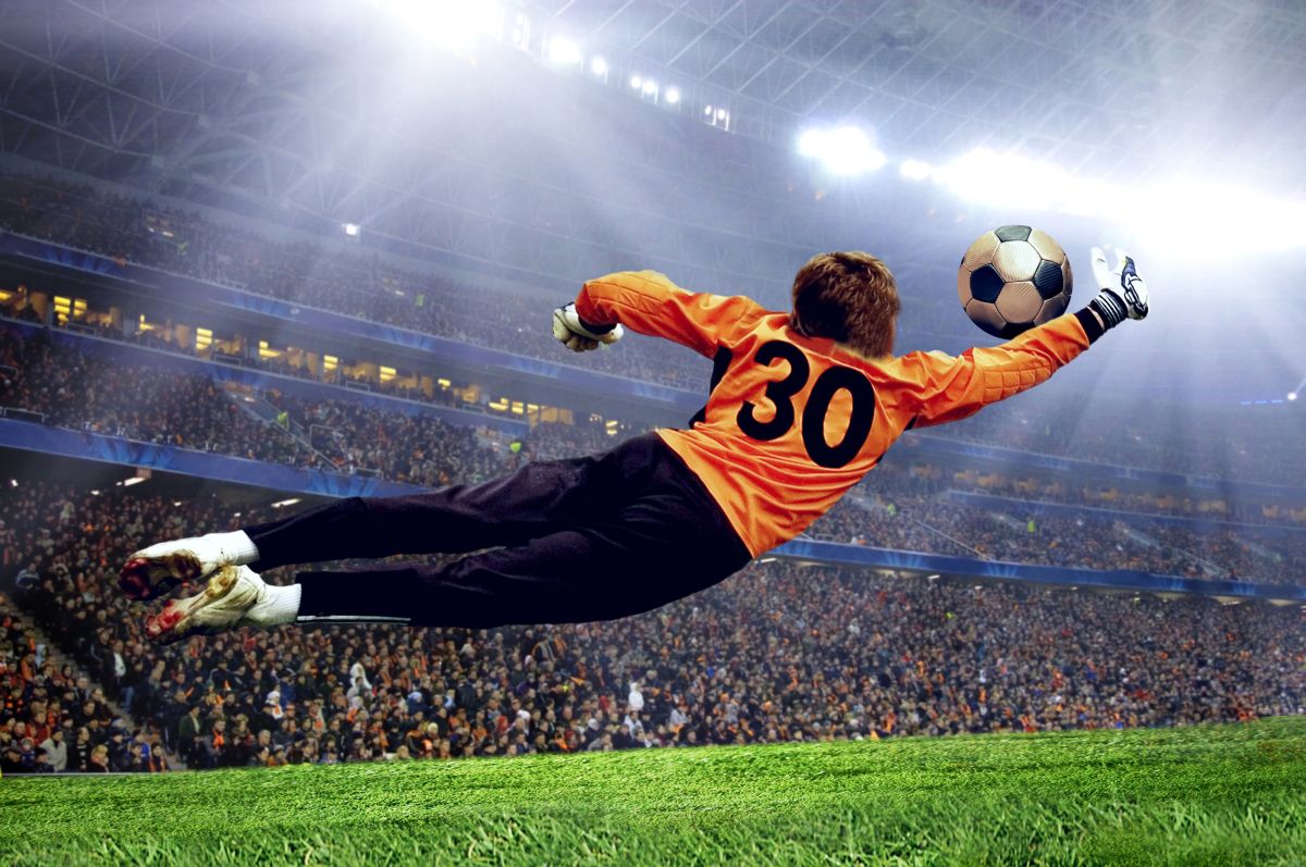 Bramkarz rzuca się w prawą stronę zatrzymując piłkę ręką na tle stadionu z reflektorami