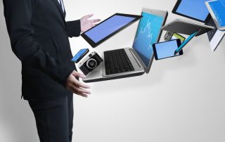 Mężczyzna w garniturze stoi bokiem po lewej obejmując rękami laptop tablet smartfon które jakby wychodzą z niego w prawy górny róg