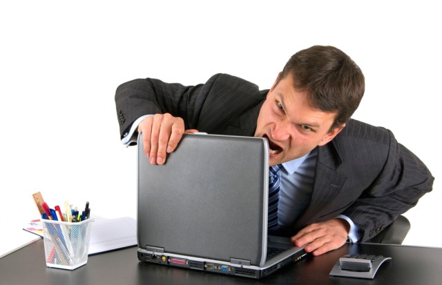 Wściekły biznesmen chce ugryźć przy biurku górną krawędź otworzonego laptopa