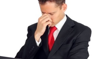 Mężczyzna w garniturze trzyma na kolanach laptop i chwyta się za nos ze spuszczoną głową